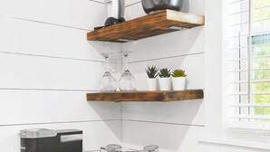 Une cuisine aux murs blancs, avec des étagères en bois, une machine à café, un évier et une fenêtre équipée de stores.