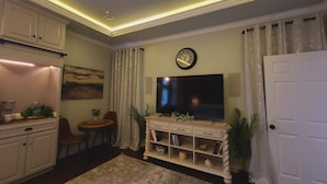 A living room with a flat-screen TV, a clock, a dining area with a table and chairs, and a kitchen area with cabinets and a countertop. - Guest Efficiency at Gold Farm (McKinney)