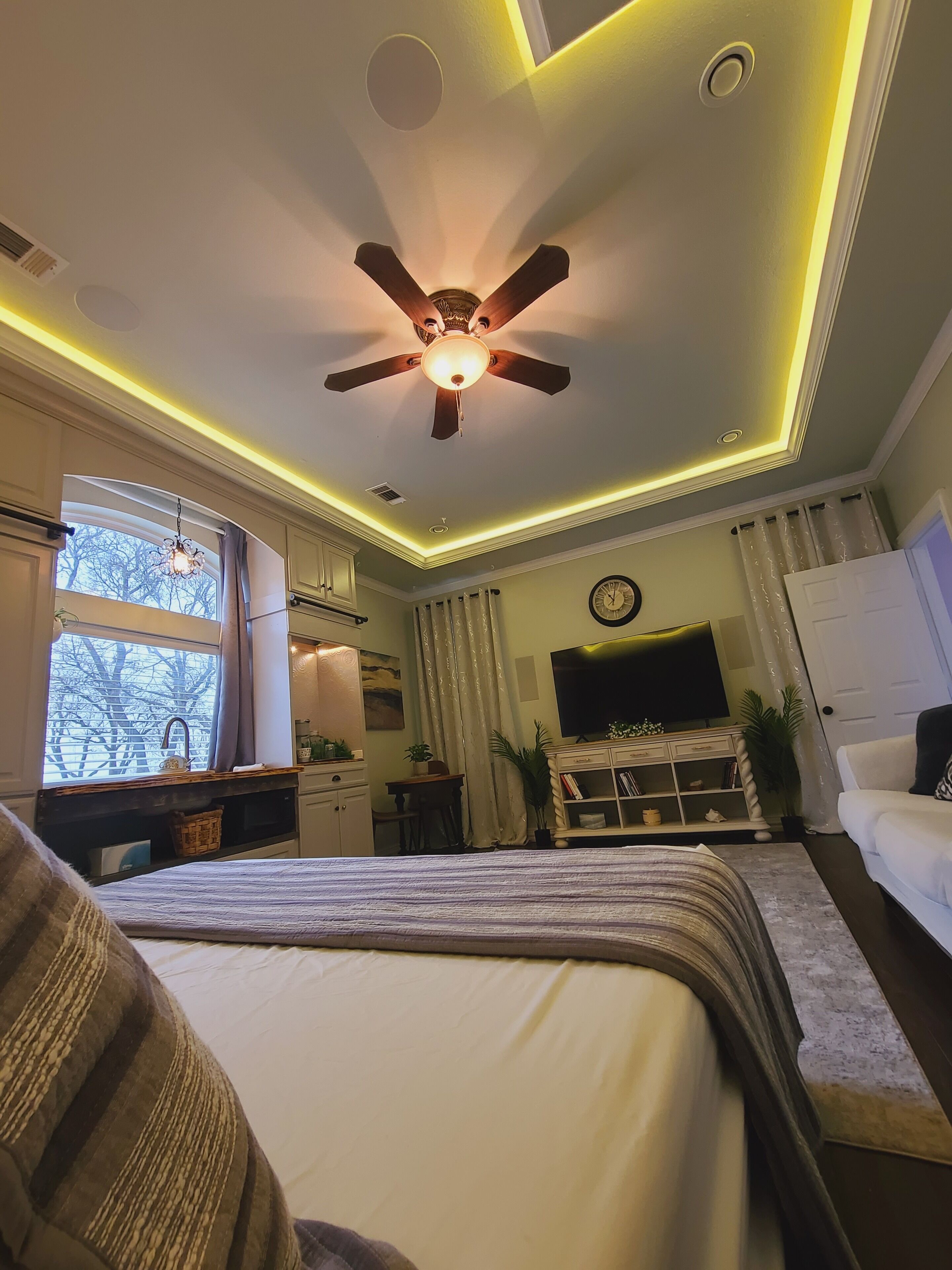 A bedroom with a bed, a ceiling fan, a TV, a desk, a chair, and a window with curtains.
