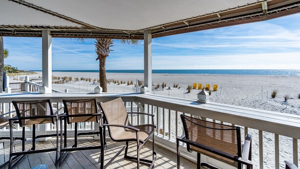 Een overdekt terras met stoelen en een bank, met uitzicht op een strand met zand en water.