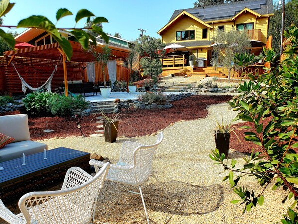 Una casa con patio, mobiliario exterior y jardín con plantas y un pequeño elemento acuático.