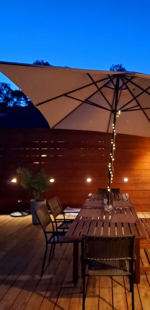 Una terraza de madera al aire libre con una mesa, sillas, un paraguas grande y luces colgantes.