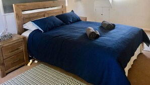 Een slaapkamer met een houten hoofdbord, een blauwe spreidek, twee opgerolde handdoeken op het bed, een kamerkleed met dessin en een nachtkastje met een zakdoosje.
