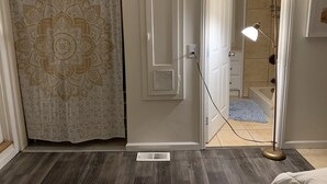Een kamer met een gordijn met een patroon, een witte deur, een ventilatie rooster, een vloerlamp en een glimp van een badkamer met een badkuip en douche.