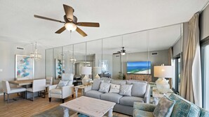 A modern living room with a ceiling fan, a dining area with a table and chairs, a sofa, and a large window with a view. - The Palms 513 by Liquid Life (Orange Beach)