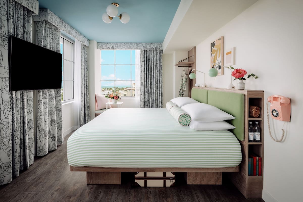 A bedroom with a large bed, a TV, a wooden headboard, a bedside table, a nightstand, a window with curtains, a wall-mounted phone, and a shelf with books and a vase.