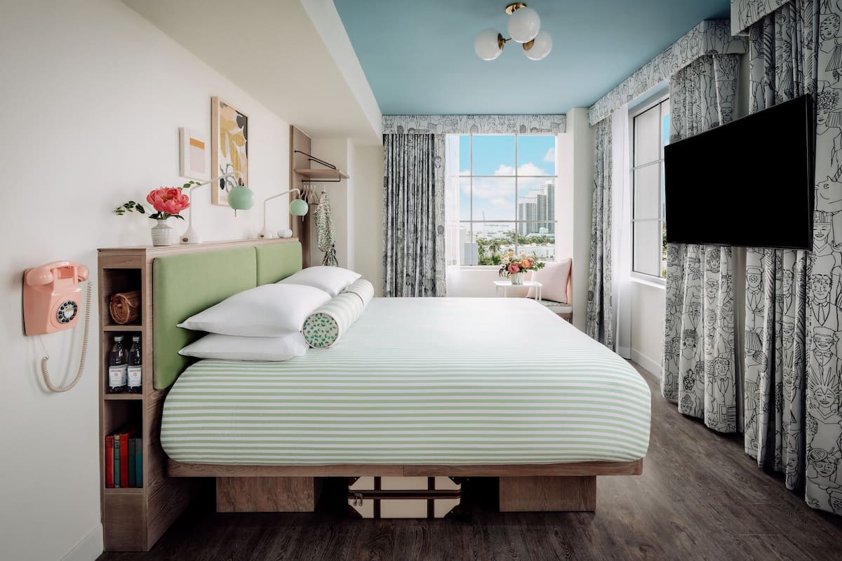 A bedroom with a bed, a bedside table, a lamp, a window with curtains, and a TV mounted on the wall.