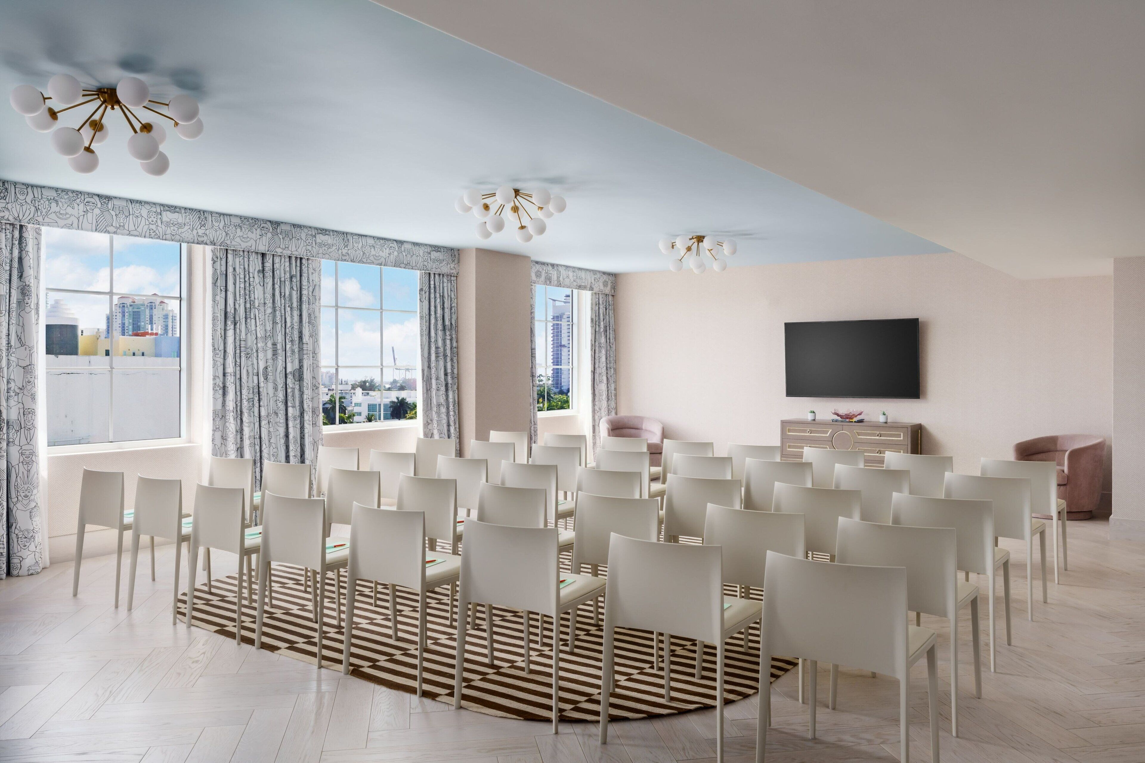 A modern conference room with white chairs arranged in rows, a large window with a view of buildings, and a flat-screen TV on the wall.