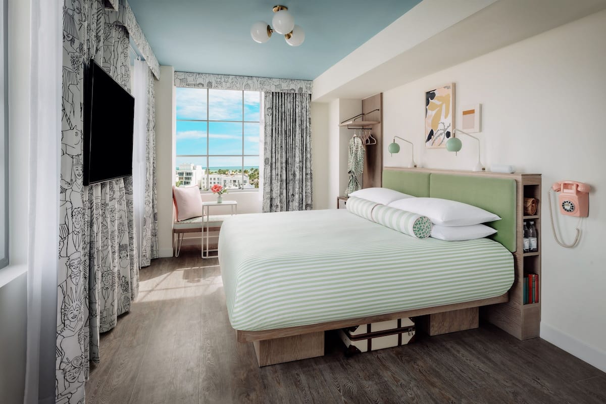 A hotel room with a bed, a desk, a chair, a window with curtains, a wall-mounted phone, and a ceiling light fixture.