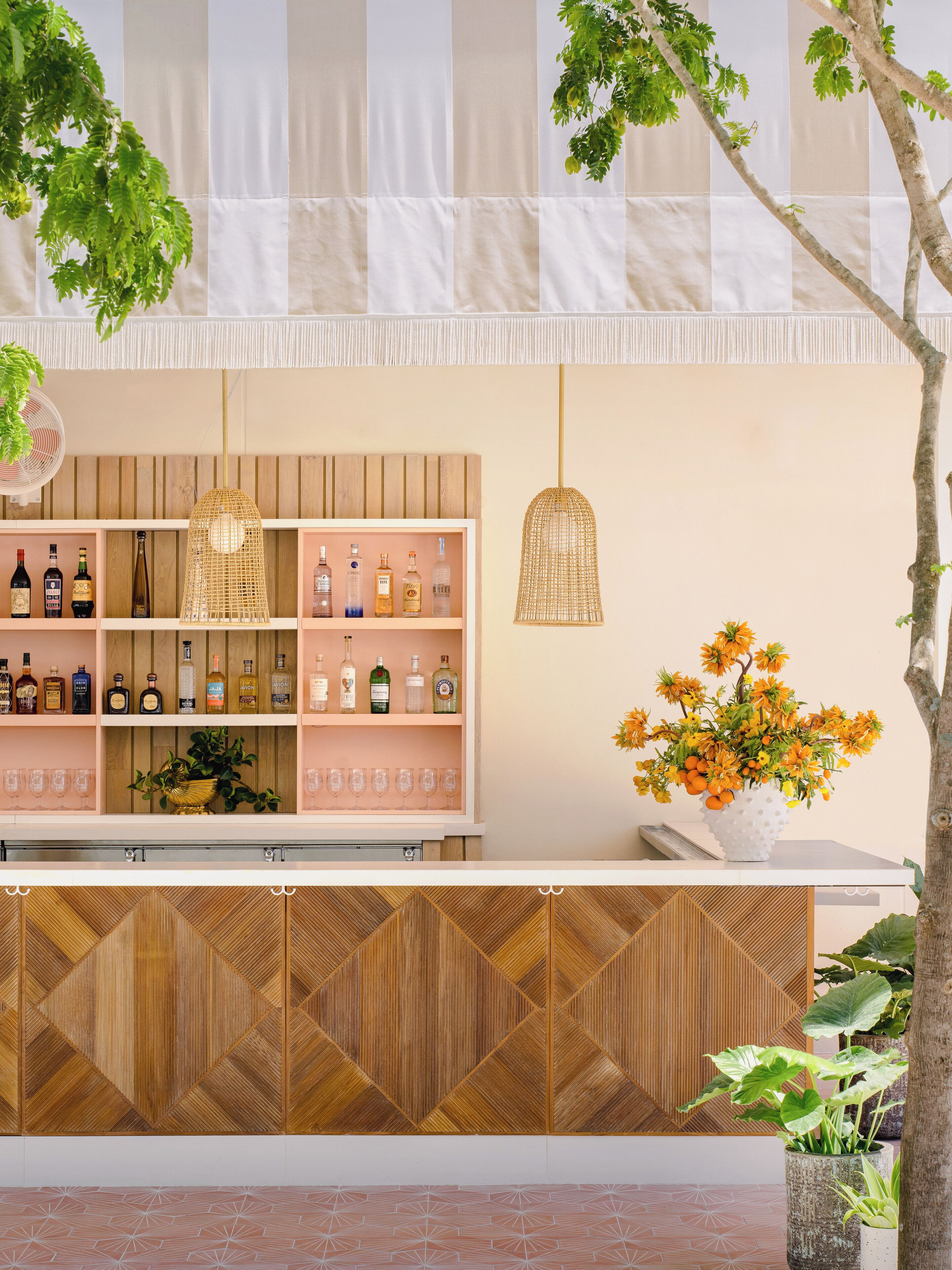 A bar area with shelves of bottles and glasses, a hanging light fixture, and a potted plant with flowers.