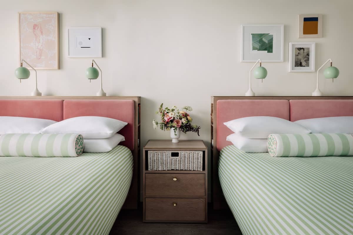 A room with two beds, each with a pink headboard and green-striped bedding. There is a nightstand with a vase of flowers and two wall-mounted lamps.