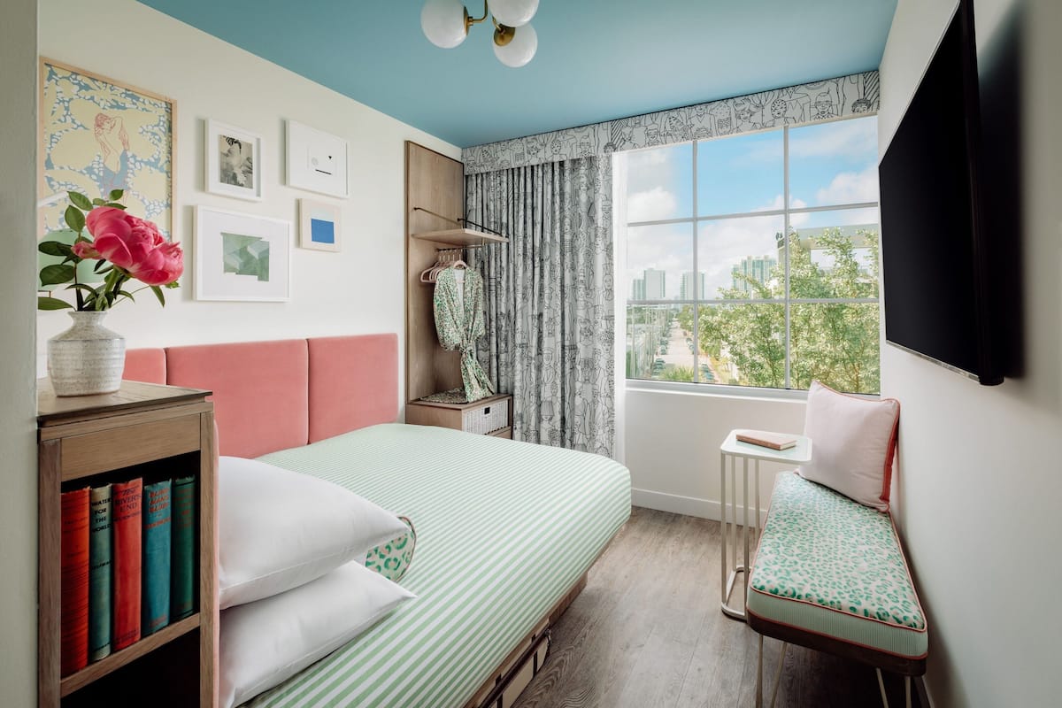 A bedroom with a bed, bedside table, books, a chair, and a window with a city view.