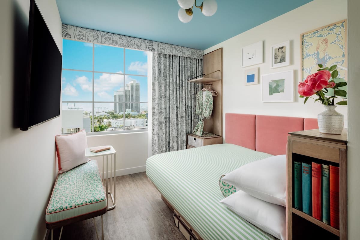 A bedroom with a large window, a bed, a bench, a bedside table with books, and a wall with framed pictures.