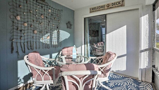 Ein sonnendurchfluteter Raum mit einem runden Tisch und Korbstühlen, eine Wand, verziert mit Muscheln und Seesternen, und ein Schild mit der Aufschrift 'WELCOME TO OUR DECK'.