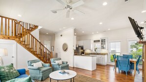 A spacious living area with a wooden staircase, a kitchen with white cabinets, and a dining area with blue chairs.