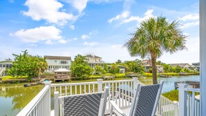 A balcony with wicker chairs overlooking a river with boats, surrounded by houses and a palm tree.