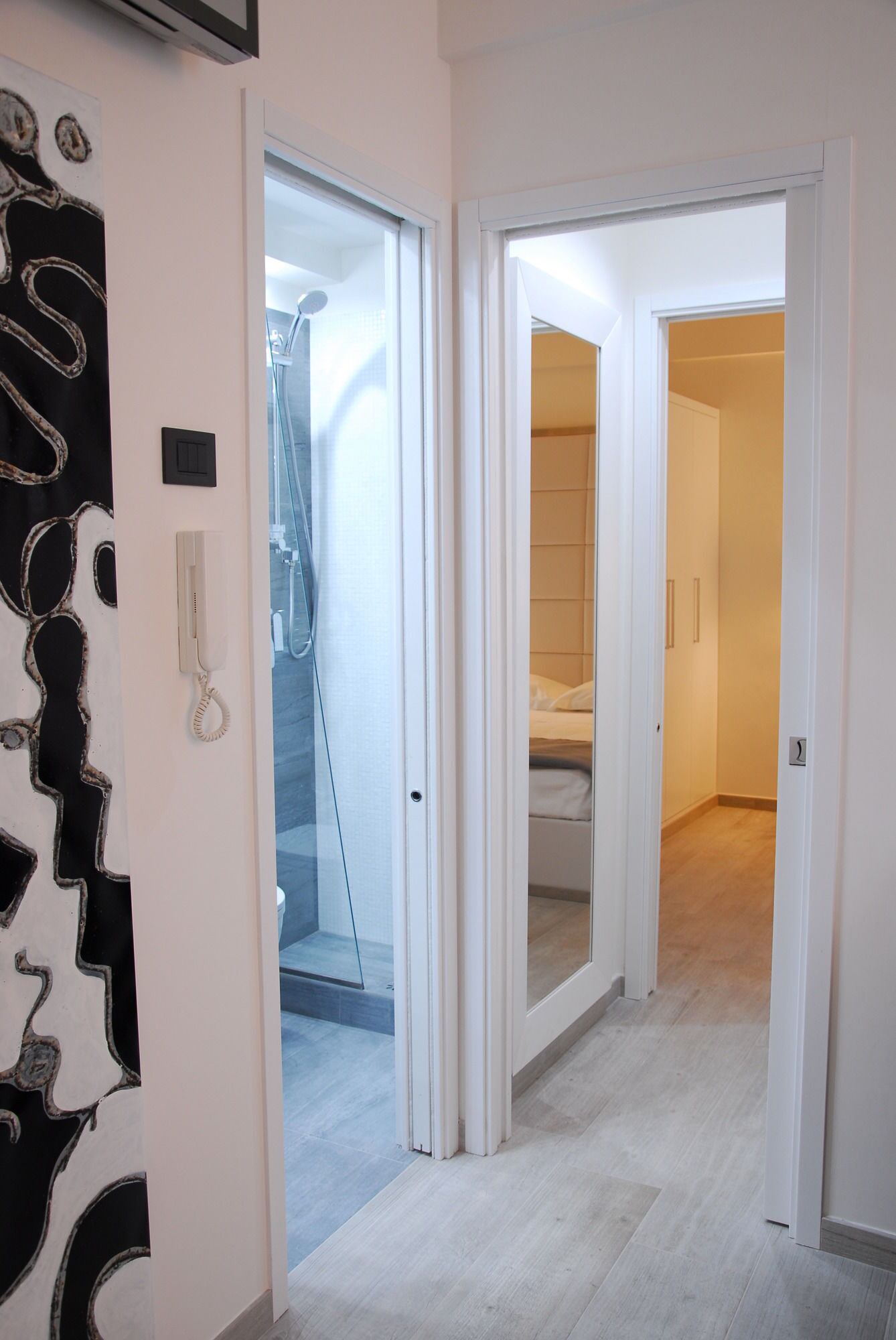 Un couloir avec une porte de salle de bain, un espace douche et une chambre à coucher visible à travers une porte ouverte.