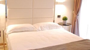 Ein ordentlich bezogenes Bett mit weißen Laken und einer grauen Tagesdecke, ein großes, gepolstertes Kopfstueck, zwei Nachttischlampen und ein kleiner Topfpflanze auf einem Beistelltisch.