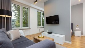 Ett modernt vardagsrum med en grå soffa, ett soffbord och en platt-TV som är monterad på väggen. Stora fönster släpper in naturligt ljus och ger utsikt över grönområden utanför.