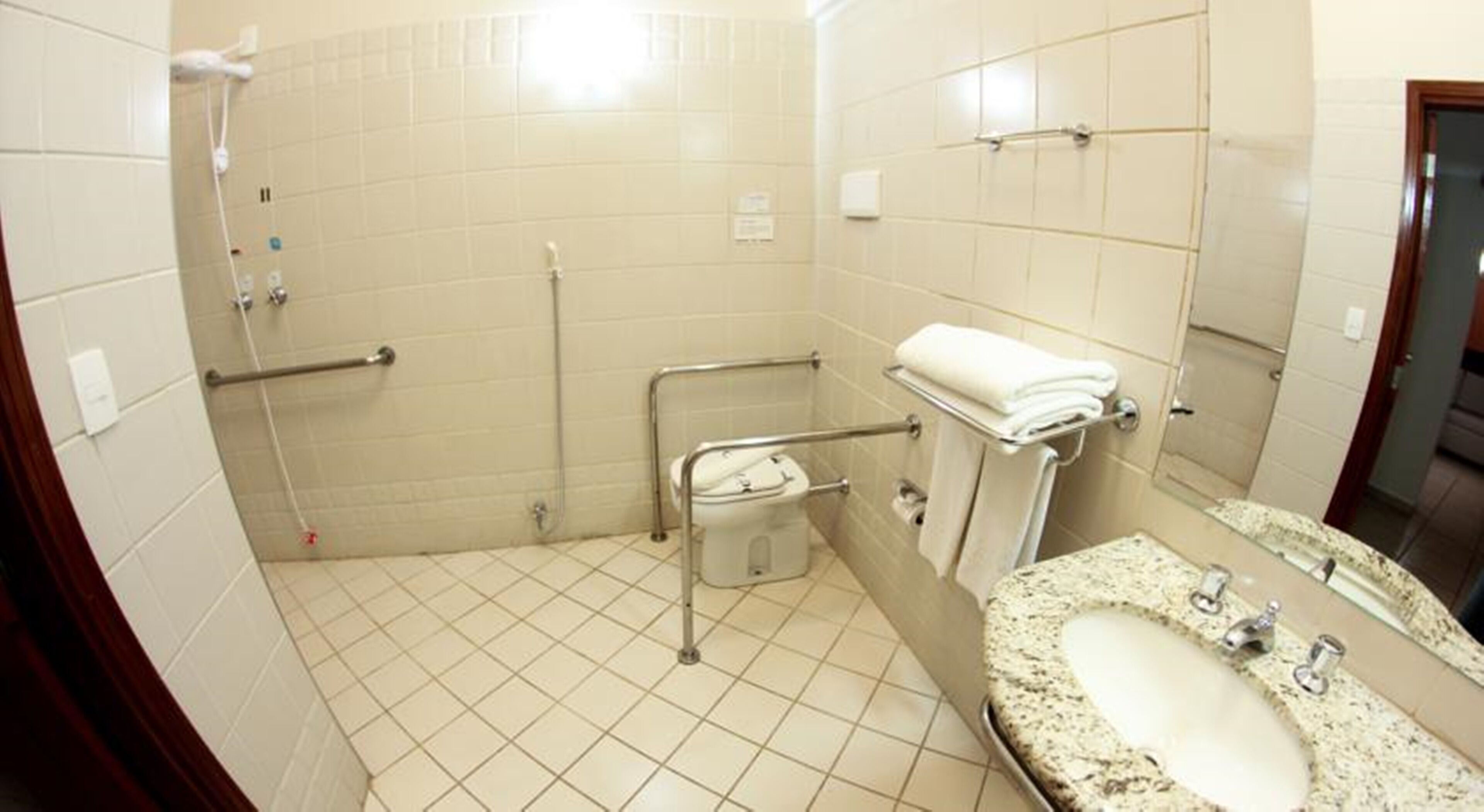Ett badrum med ett kaklat golv, ett handfat med en bänkskiva i granit, en toalett med ett stödhandtag och en duschhörna med ett duschmunstycke som kan hållas i handen.