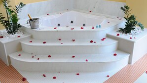 A corner bathtub with a champagne bottle and glasses, surrounded by white steps and red rose petals. - Ibis Styles Rio Preto Monte Libano (São José do Rio Preto)
