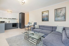 A modern living room with a grey sofa, a glass coffee table, and a dining area with a table and chairs. There are kitchen cabinets and a microwave visible in the background.
