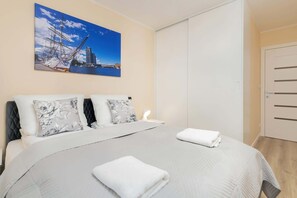 A neatly made bed with a quilted cover, two pillows with floral patterns, and a folded white towel. A painting of a bridge and cityscape is on the wall above the bed.