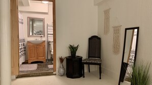 Un pasillo con una puerta de madera que da a un baño, una silla con un cojín estampado, una planta en maceta y un espejo en la pared.