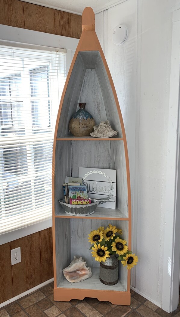 A tall, narrow wooden shelf with various decorative items, including a vase with sunflowers, a bowl with books, and a decorative piece with the text "Treasure each day". - Little cottage near the sea! Golf CART is included! (Garden City)