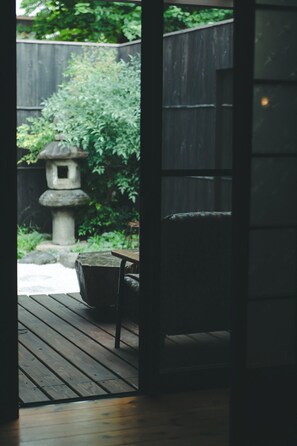 Una terraza de madera con un farol de piedra y un banco, que conduce a un jardín con valla de madera y vegetación.