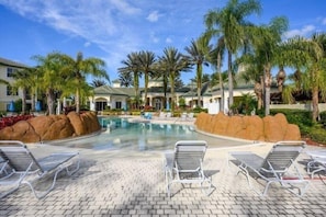 A pool area with lounge chairs, palm trees, and a building with a tiled roof.