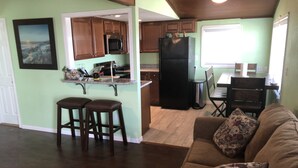 A kitchen with wooden cabinets, a black refrigerator, and a dining area with a table and chairs. There are two bar stools near the kitchen counter. - Getaway By The Bay Unit 1 (Sargent)