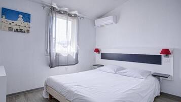 A bedroom with a bed, a window with a sheer curtain, a wall-mounted air conditioner, and a painting on the wall.