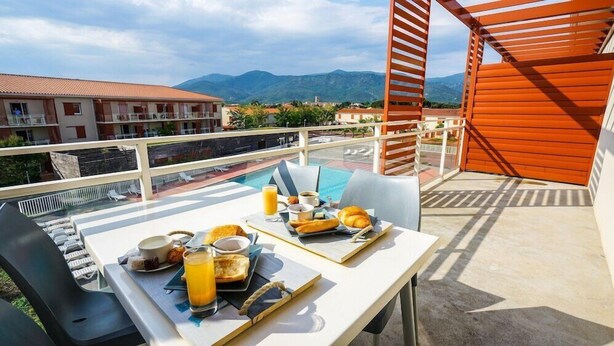 Un balcón con una mesa lista para el desayuno, con vistas a una piscina y montañas.