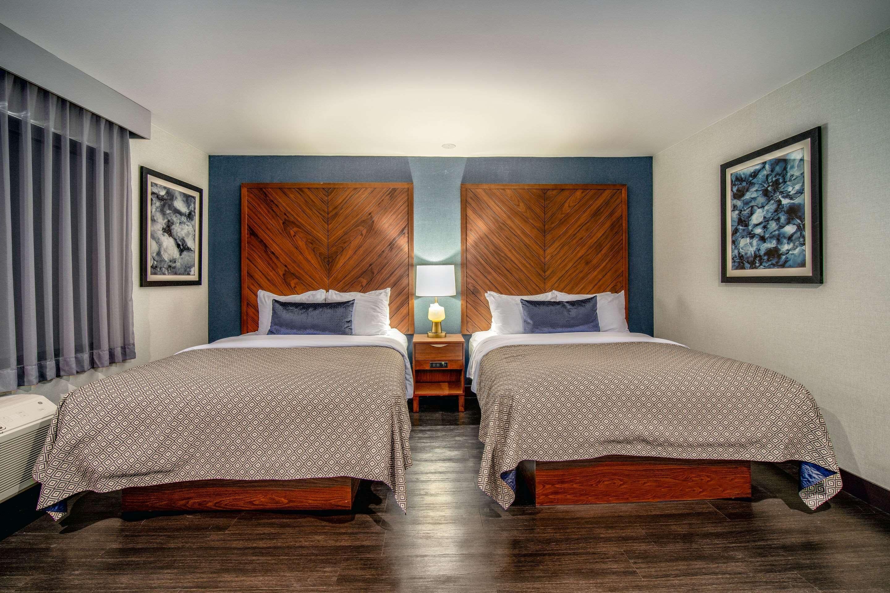 A hotel room with two beds, a wooden headboard, a nightstand with a lamp, and artwork on the walls.