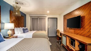 Habitación de hotel con dos camas, un televisor montado en la pared, un escritorio con lámpara y un mueble de entretenimiento de madera.
