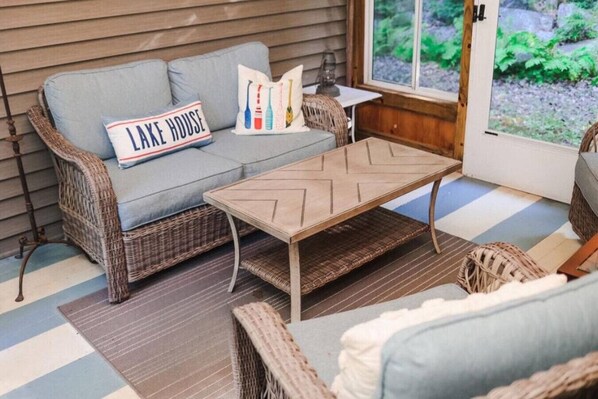 A wicker sofa with a cushion, a wooden coffee table, and a pillow with the text "LAKE HOUSE".