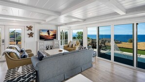 A spacious living room with a large gray sofa, a white coffee table, and a TV. There are multiple windows offering a view of the ocean. - NEW! Ocean View Hawaii Vacation And Event Retreat (Kapaau)
