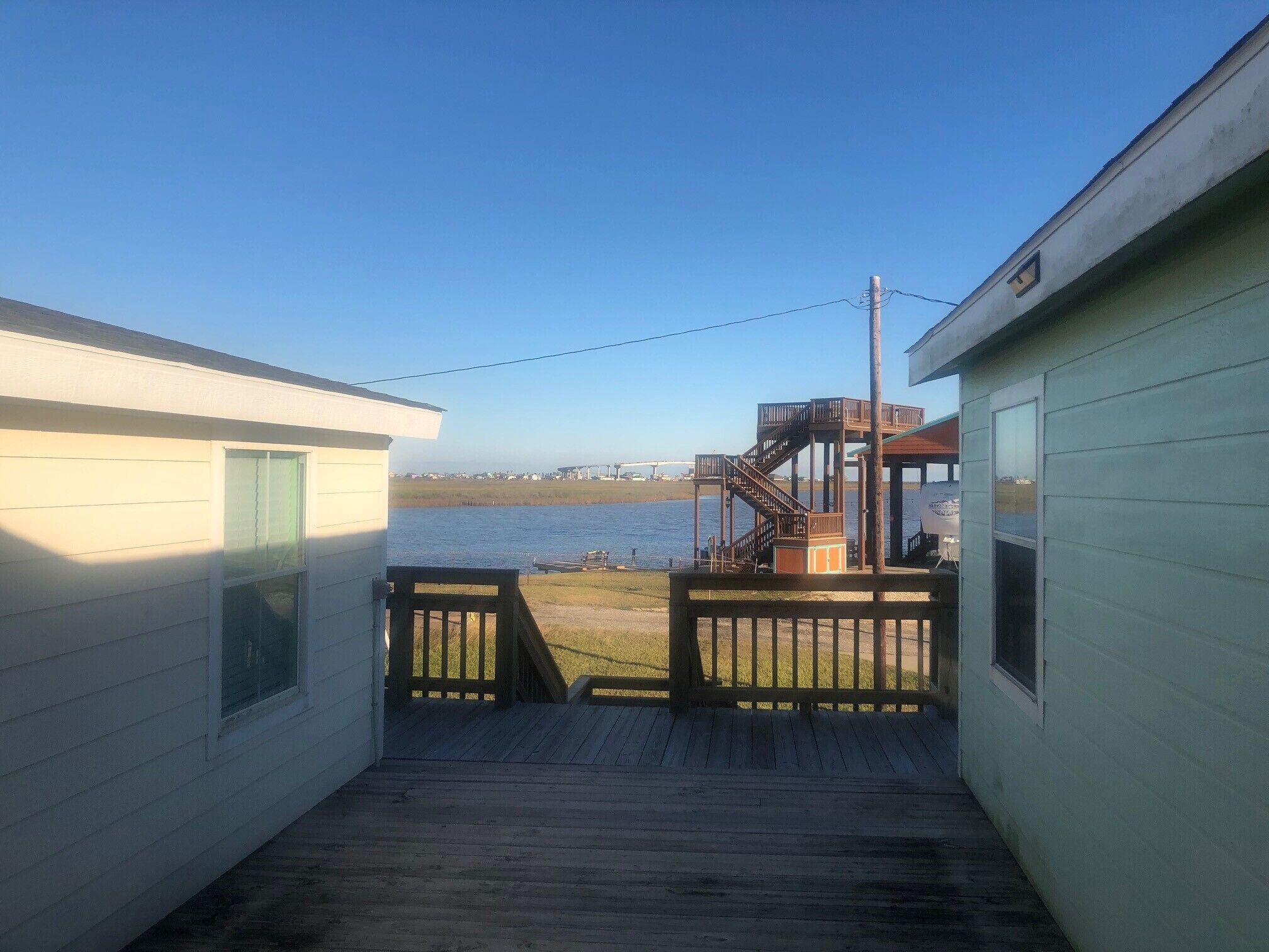 A narrow wooden deck leading to a small building with a view of a body of water and a bridge in the distance.