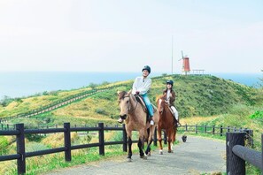 Two people riding horses on a paved path with a lighthouse in the background.