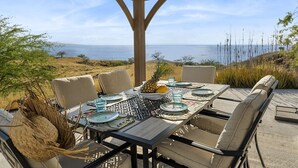 An outdoor dining area with a view of the sea, a covered pergola, and a table set with plates, glasses, and a pineapple.