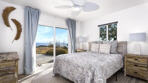 A bedroom with a large bed, a ceiling fan, a window with a view of the outdoors, and a wooden nightstand with a lamp.