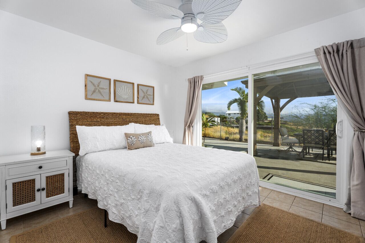 Une chambre dotée d’un grand lit, d’un ventilateur de plafond, d’une table de chevet et offrant une vue sur l’extérieur grâce à une porte-fenêtre coulissante.