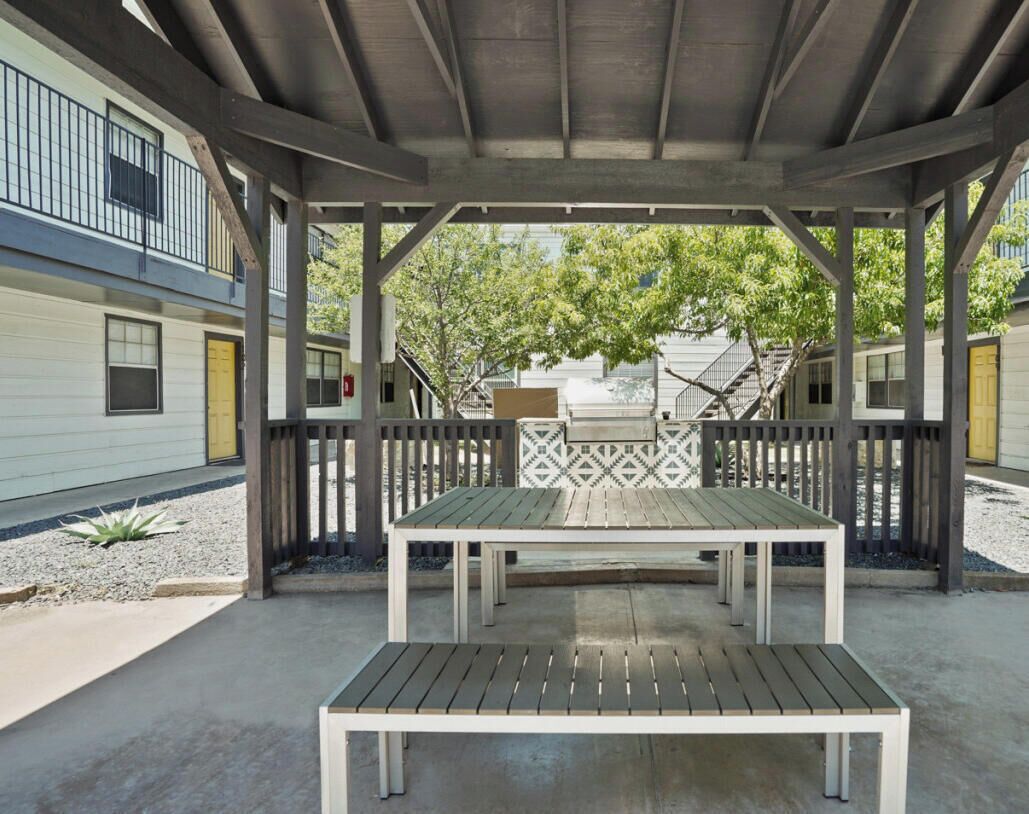 A covered outdoor seating area with a table and benches, surrounded by a wooden fence, in front of a building with balconies and windows.