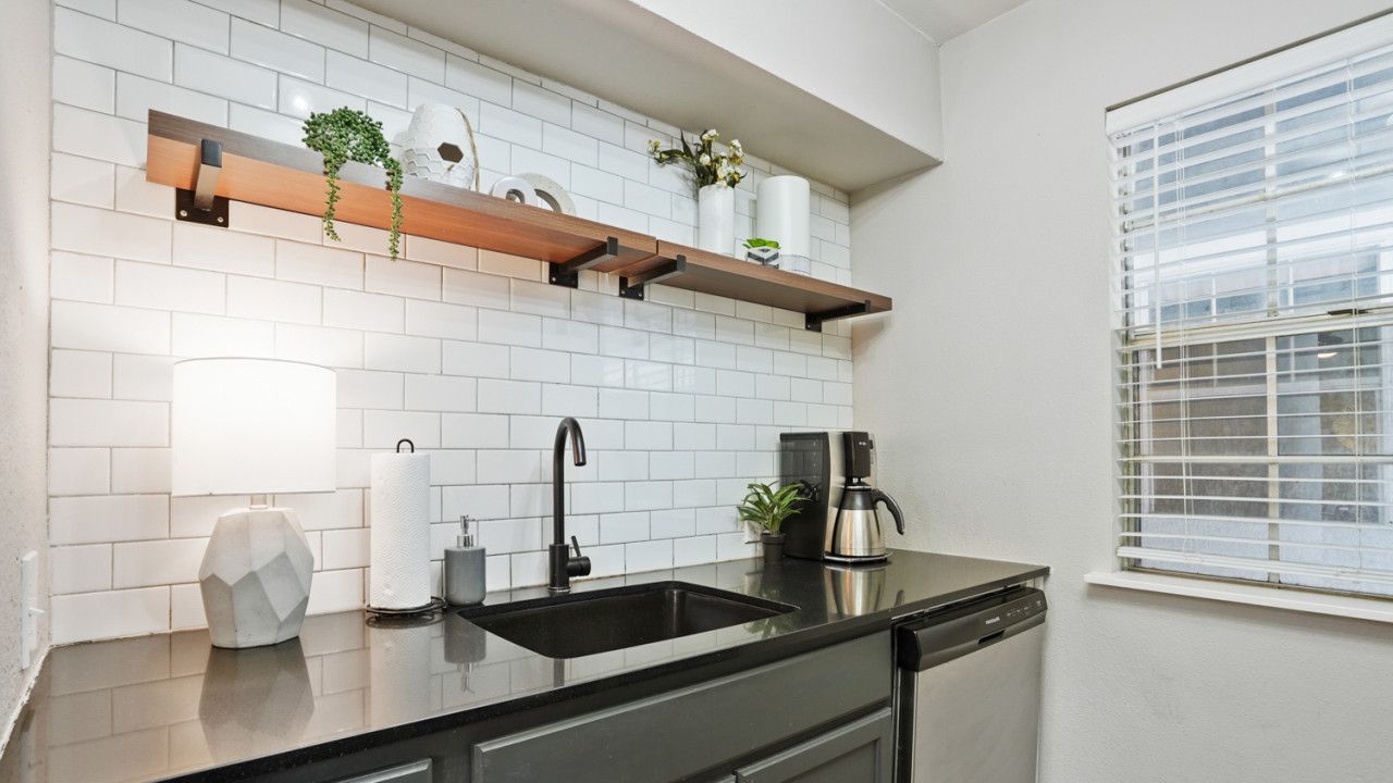Une cuisine moderne avec des carreaux de métro blancs, un plan de travail noir, un lave-vaisselle encastré et des étagères en bois ornées d’objets décoratifs.