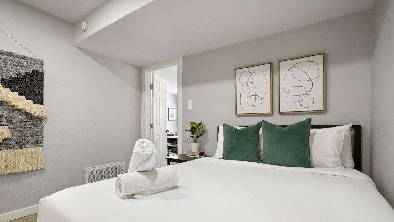 A modern hotel room with a large bed, two green pillows, a white blanket, and a wall-mounted abstract art piece.