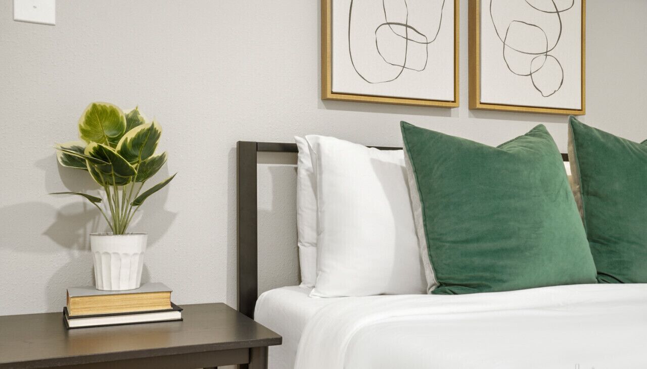 Una cama bien hecha con almohadas blancas y verdes, una mesita de noche con una planta y libros, y arte abstracto en la pared.