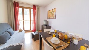 Een gezellige woonkamer met een bank, een ontbijt gedekte eettafel, een televisie en een balkon met rode gordijnen.