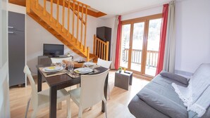 A modern living room with a staircase, a dining area with a table set for two, and a large window with curtains. - Appartement Duplex Au Pied des Pistes (Les Orres)