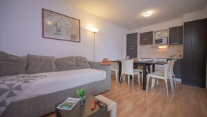 A modern living room with a grey sofa, a dining table with chairs, a kitchen area with dark cabinets and a microwave, and a framed picture on the wall. - Appartement avec Balcon Privé  | Piscine (Les Orres)